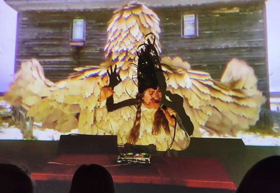 Gjennom direktestrømming fra Moskva, framførte Podkorytova en performance kalt «Global Silent Spell» i Bodø. Her synger hun til menn og ber dem komme tilbake fra krigen. Filmen i bakgrunnen er fra landsbyen Nenoksa utenfor Arkhangelsk, innspilt noen måneder etter en atomulykke ved en militærbase der. (Foto: Prosjektet Border / No border)